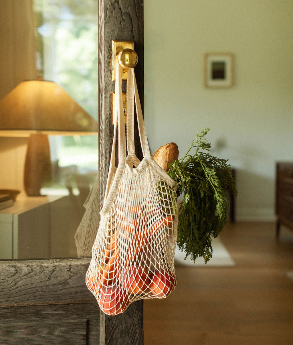 A cream mesh grocery bag hanging from a brass hook on a rustic wooden door handle, filled with produce and herbs in a softly lit home interior.